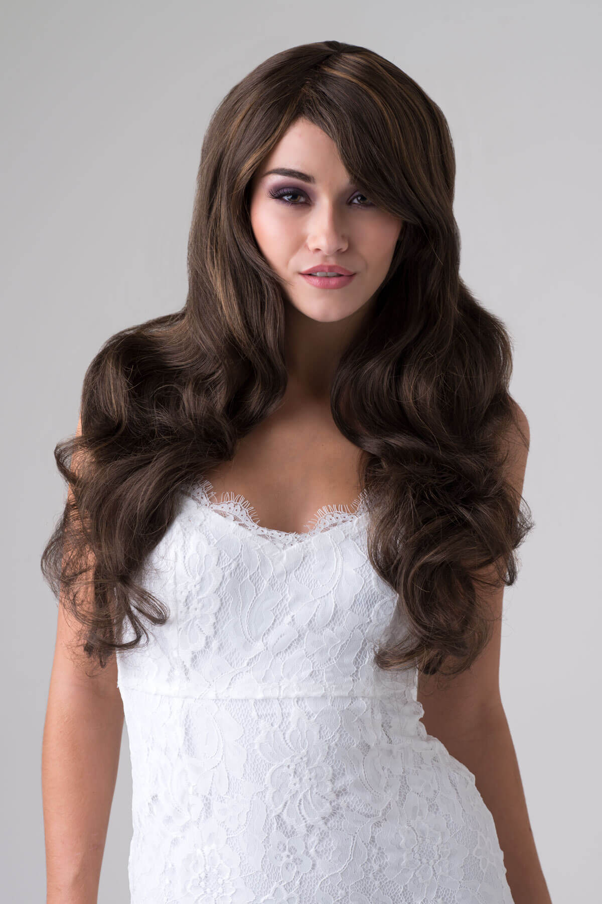 Woman wearing a long brown wig with a white lace dress on a grey background. Long brown wig with gentle waves: Bella brown with blonde highlights 8H27