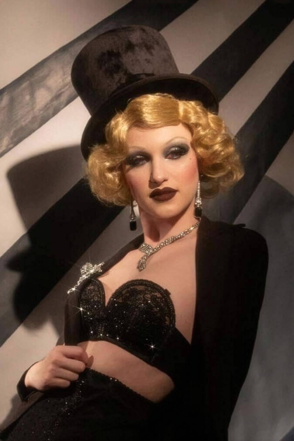 Person in drag with blonde wig, top hat, and dark makeup against a striped background. Blonde 1920s 1930s style wig, short with finger waves: Diva warm blonde 24B
