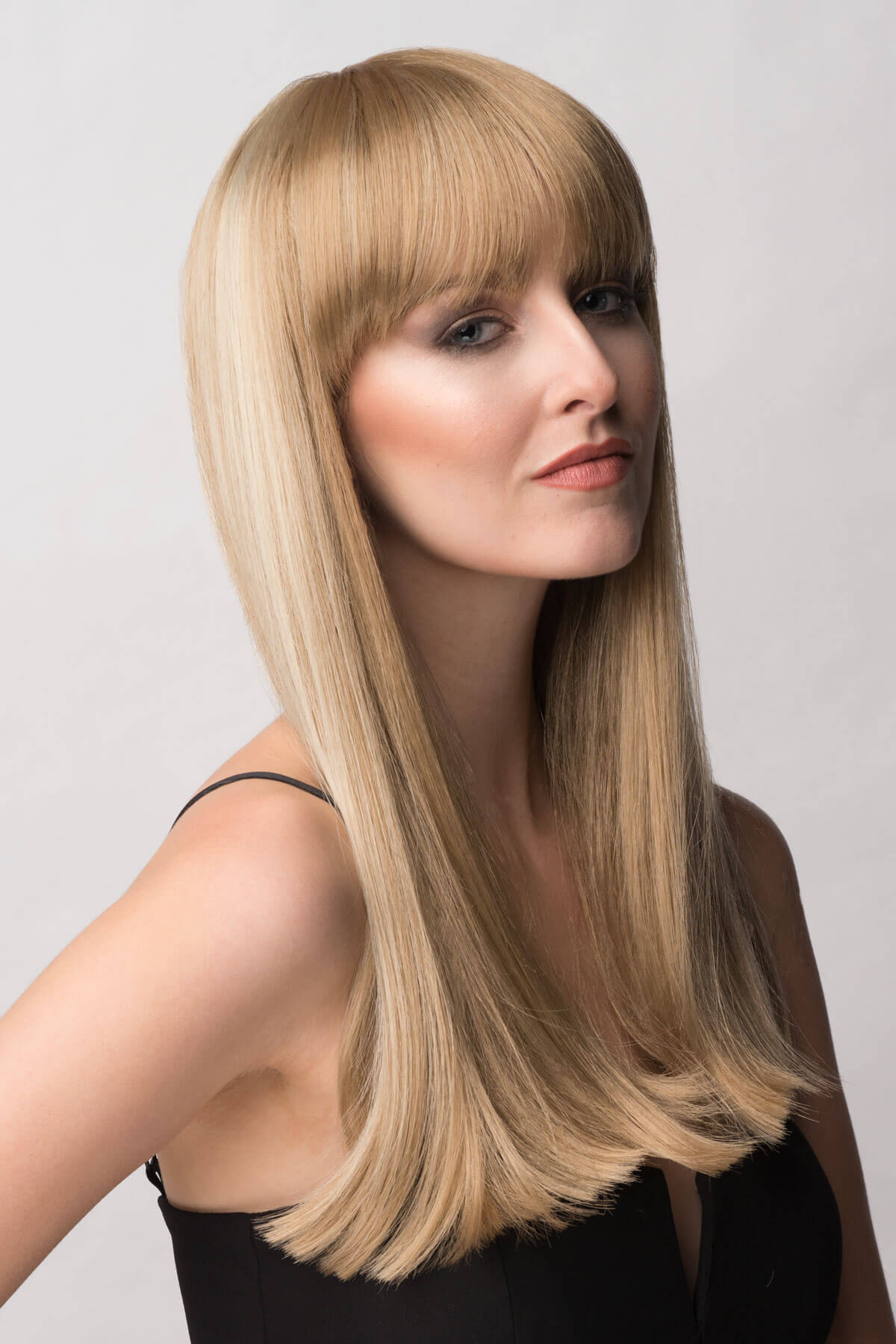 Woman with long blonde hair styled with bangs against a plain background. Long blonde wig with Highlights and straight fringe: Tilly