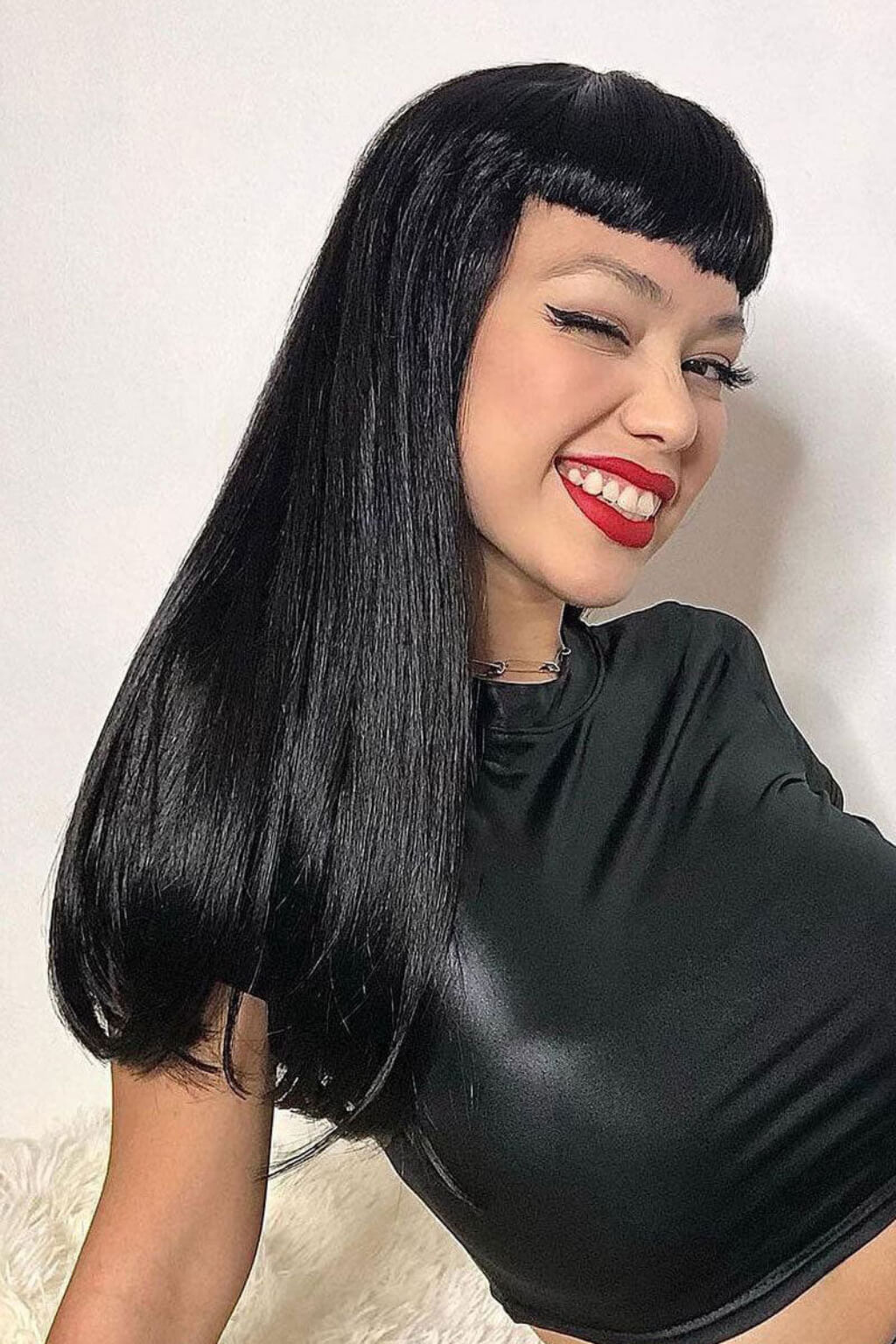 A woman wearing a long, straight black wig with blunt bangs, styled with red lipstick and a black crop top against a plain light background. Long black wig with straight hair and a short, straight fringe: Maxine