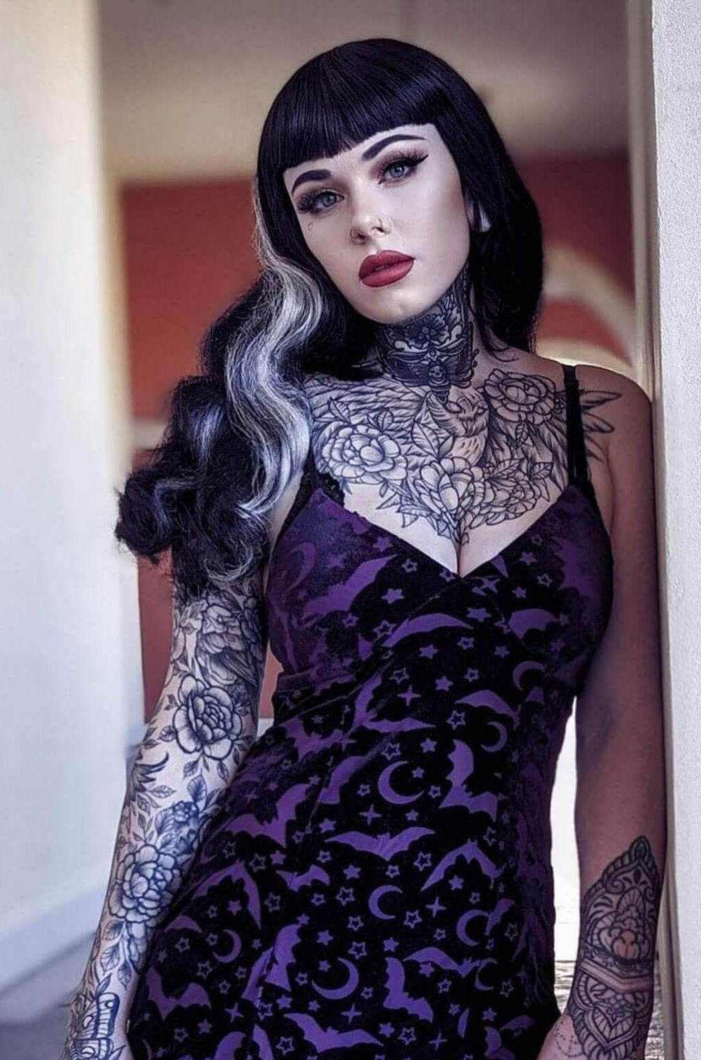 Woman wearing a purple lace dress with tattoos on her arms and neck. Long black 1950s pin up style wig with platinum streak and retro fringe: Michelle