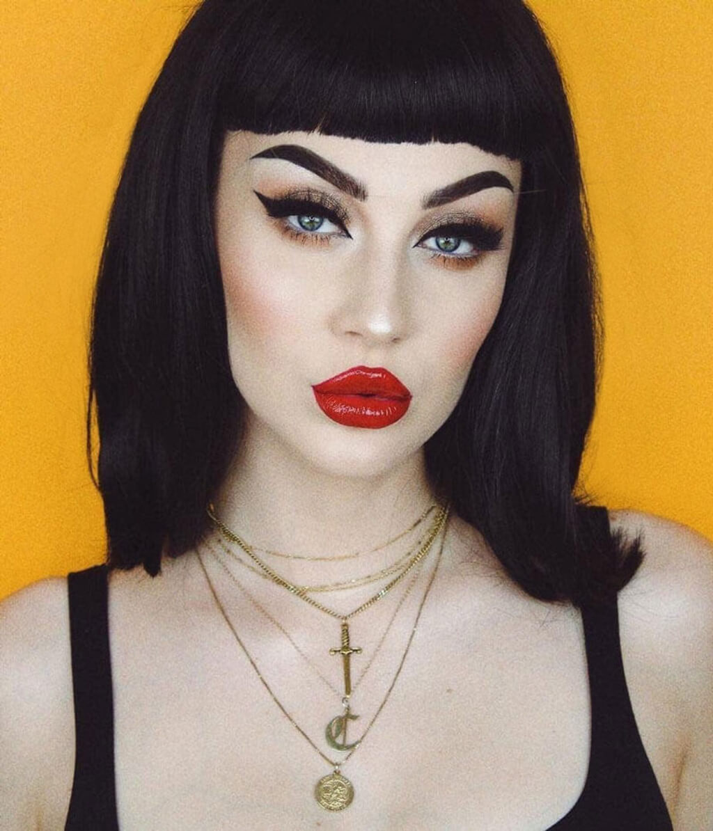 Woman with bold makeup and black bob haircut against a yellow background. Black long bob wig with a short retro-style fringe: Beth