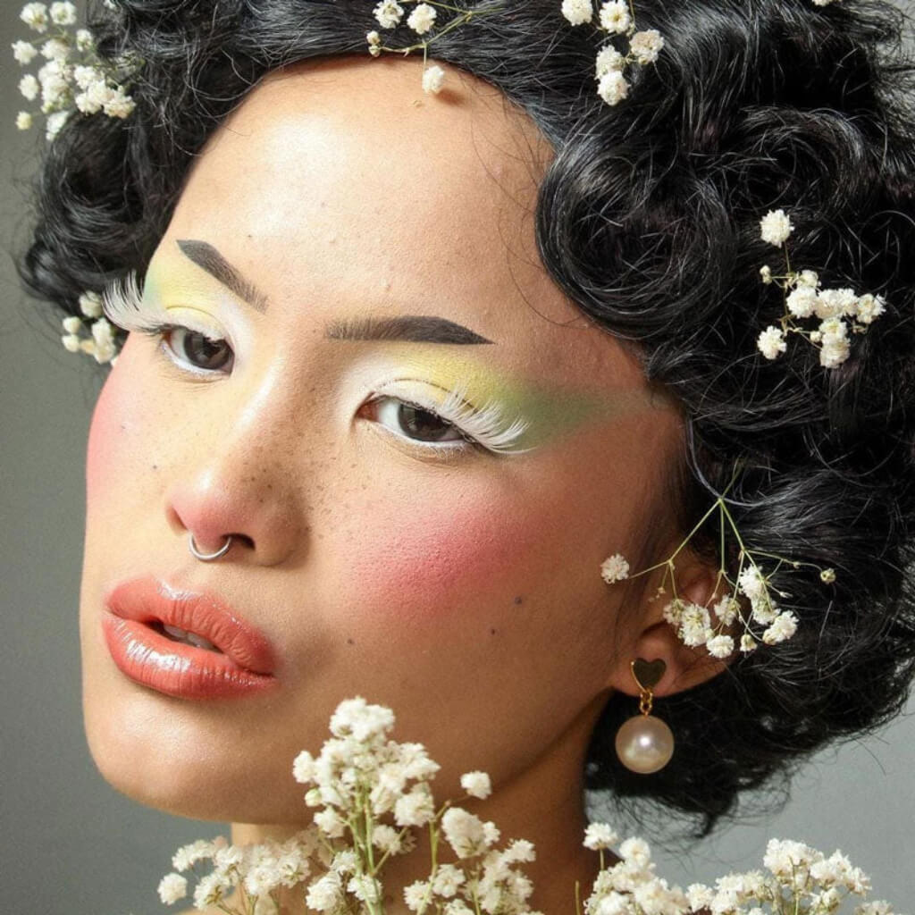 Woman with floral hair and makeup, wearing pearl earrings, against a neutral background. Brown 1920s 1930s style wig, short retro style with finger waves: Cecelia dark brown 4