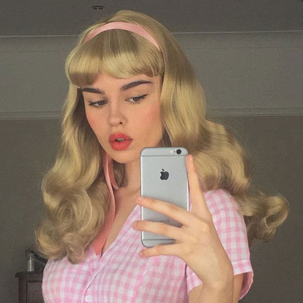 Person with vintage-style blonde hair and pink headband taking a mirror selfie.