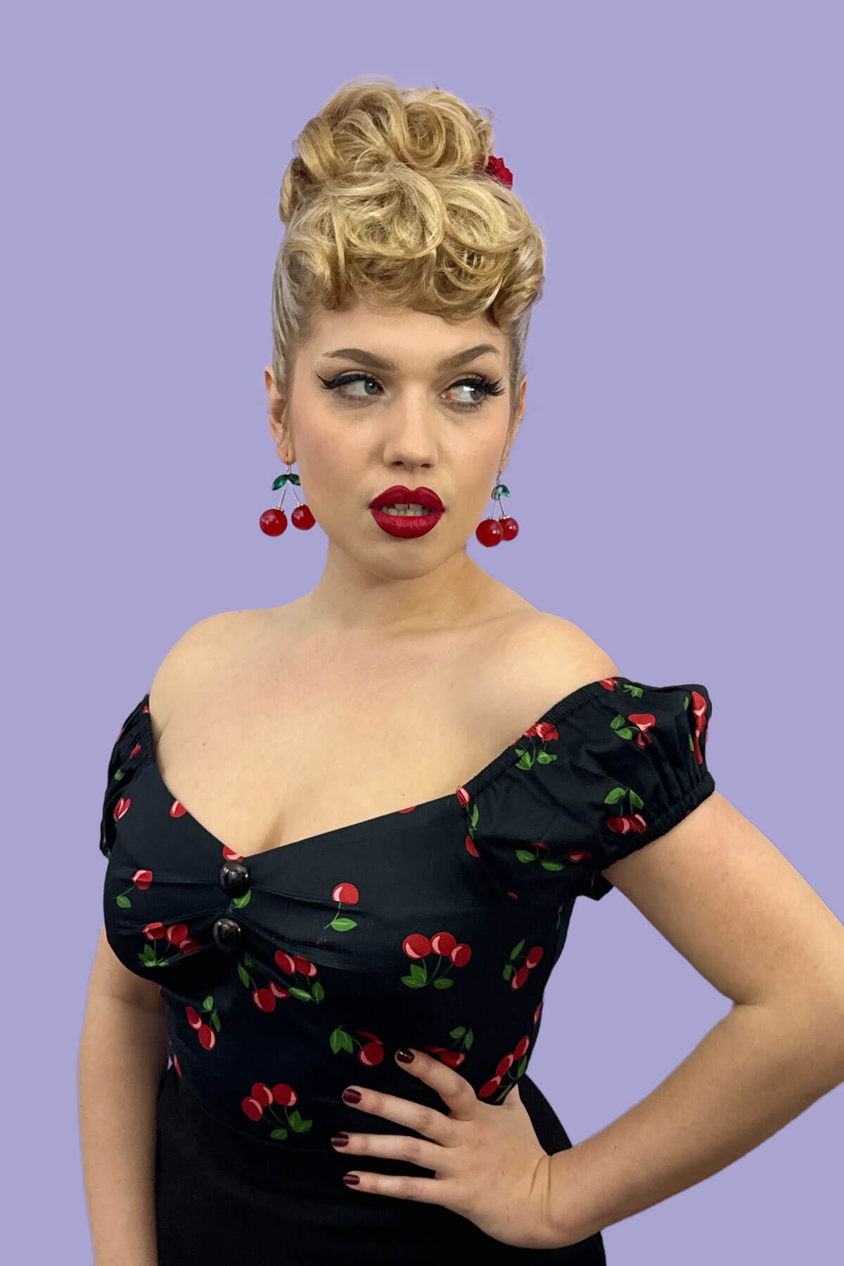 Woman wearing a black dress with red cherry patterns against a purple background. Curly bun hairpiece, rockabilly style: Toni blonde 613T24