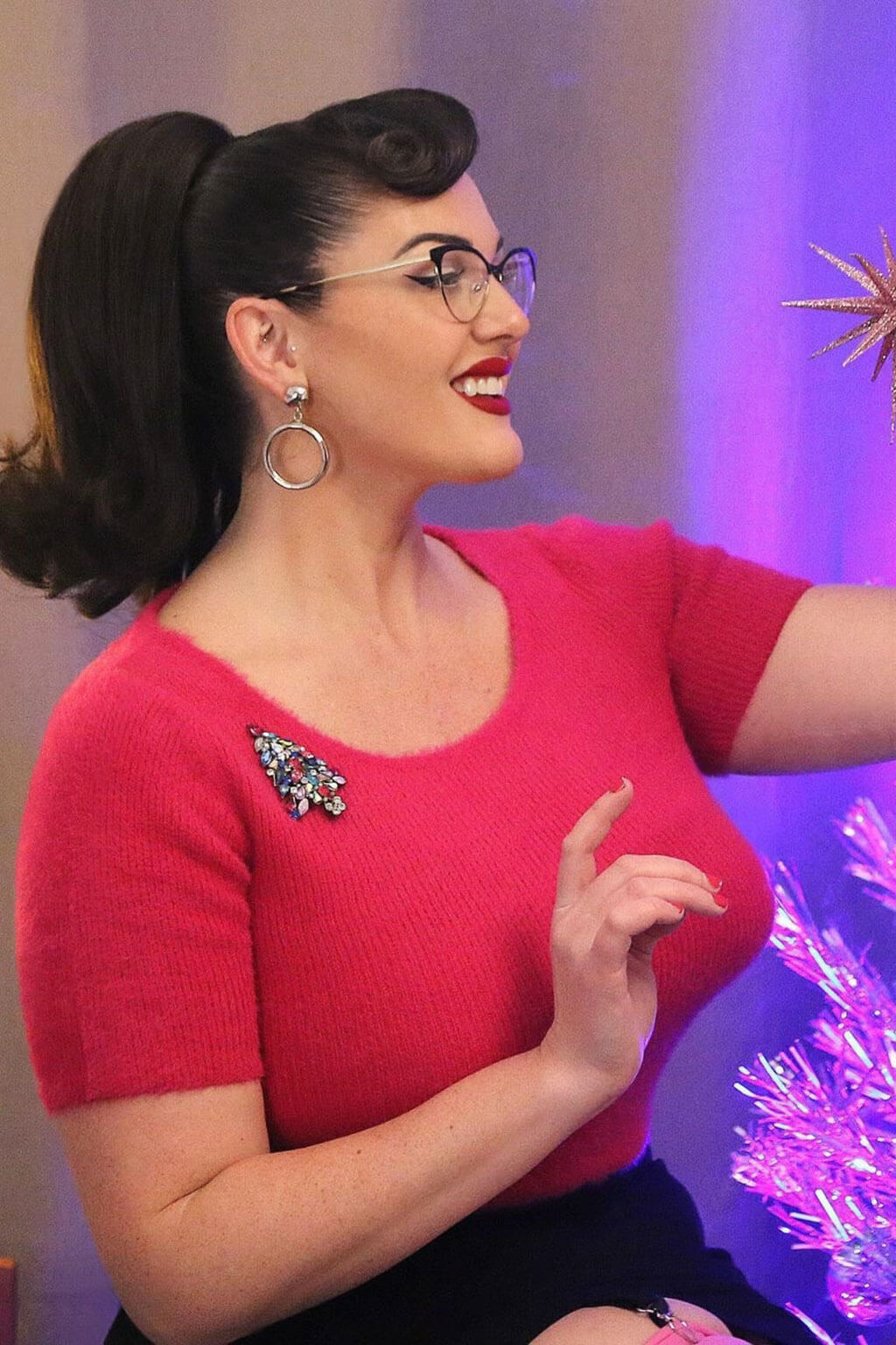 Woman in a red top with a brooch, wearing glasses and earrings, against a colourful background. Ponytail hairpiece, shoulder length with flick/flip, retro 1960s style: Ariana dark chocolate brown 6