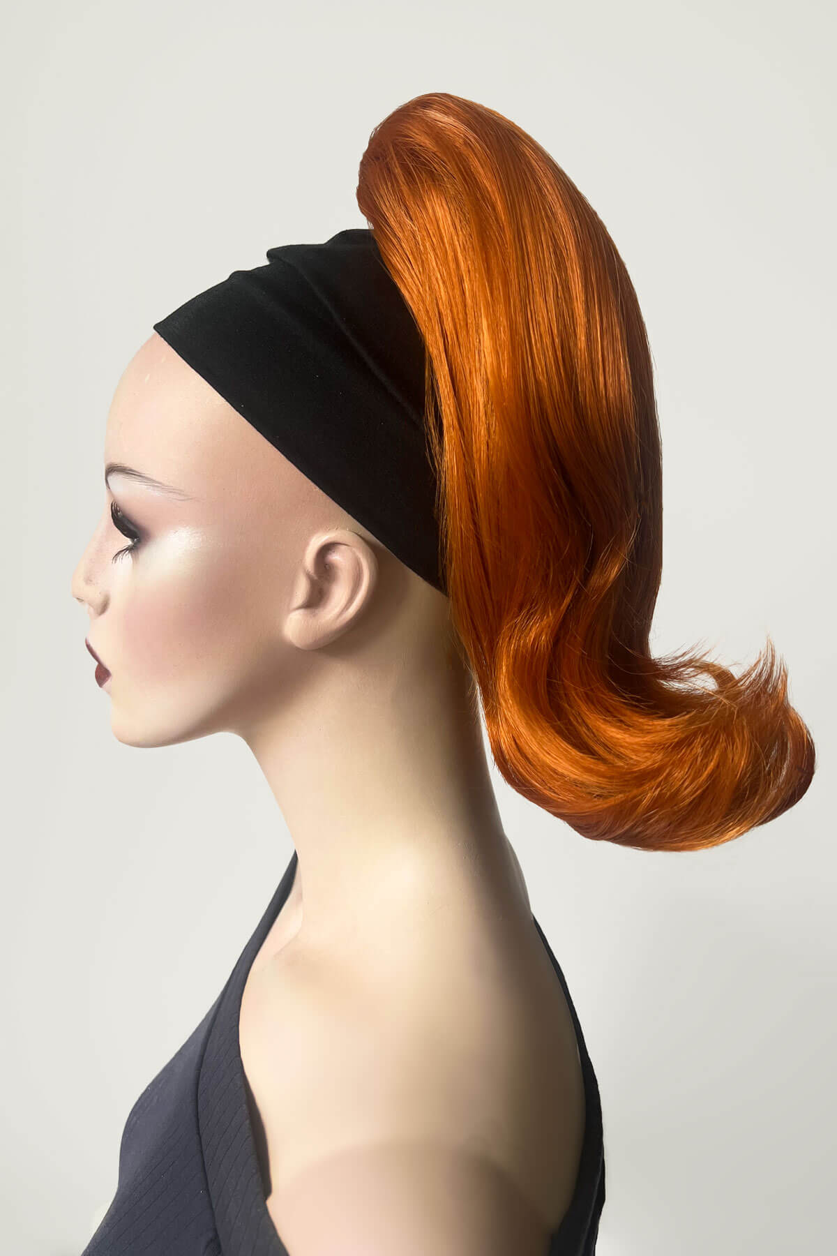 Mannequin head with orange hair styled in a ponytail against a plain background. Ponytail hairpiece, shoulder length with flick/flip, retro 1960s style: Ariana Ginger