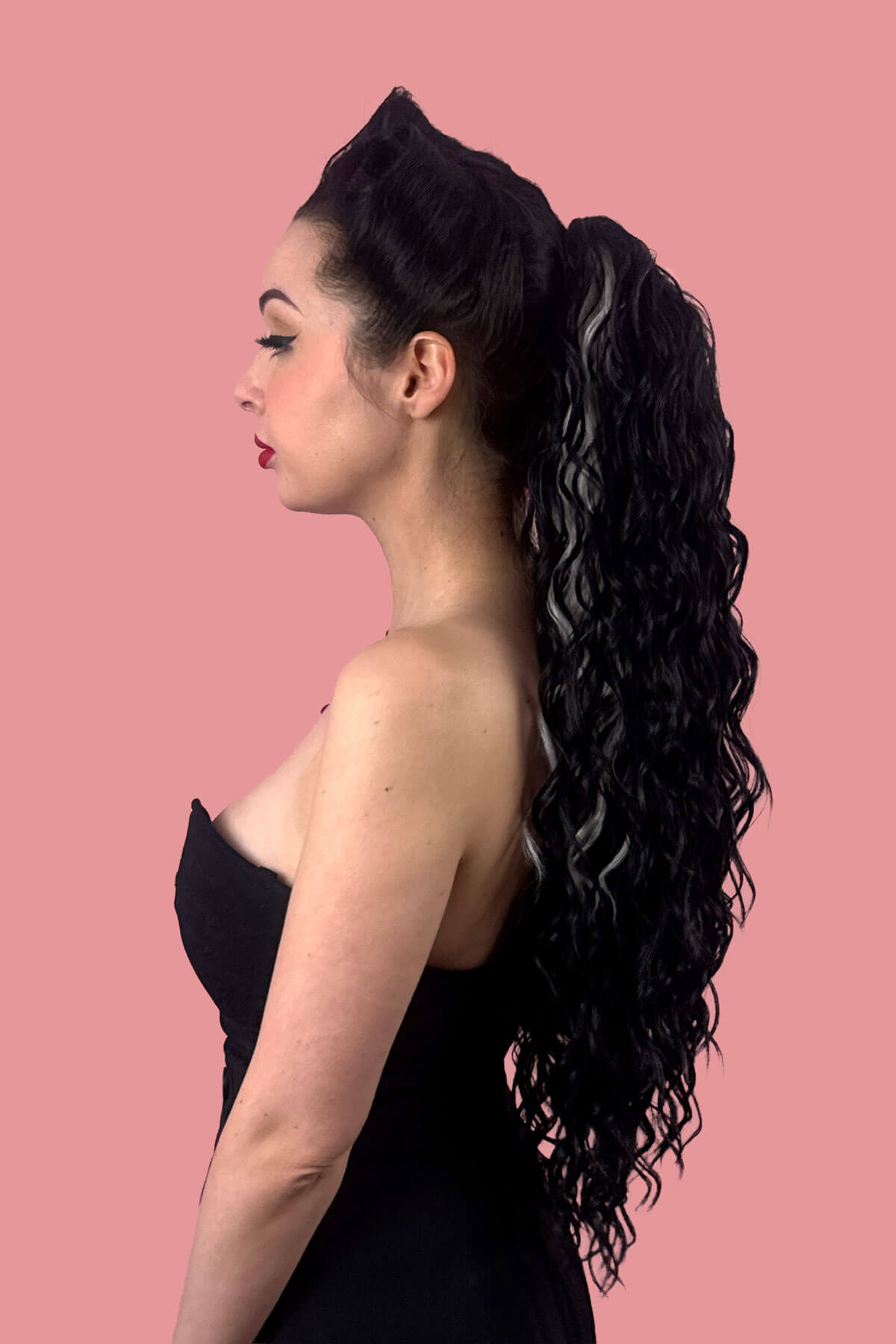 A woman in profile wearing a black strapless dress, showcasing a long, dark, curly wavy hair extension with grey streaks against a solid pink background. Long Curly Ponytail Hair Piece Extension, Black and Grey