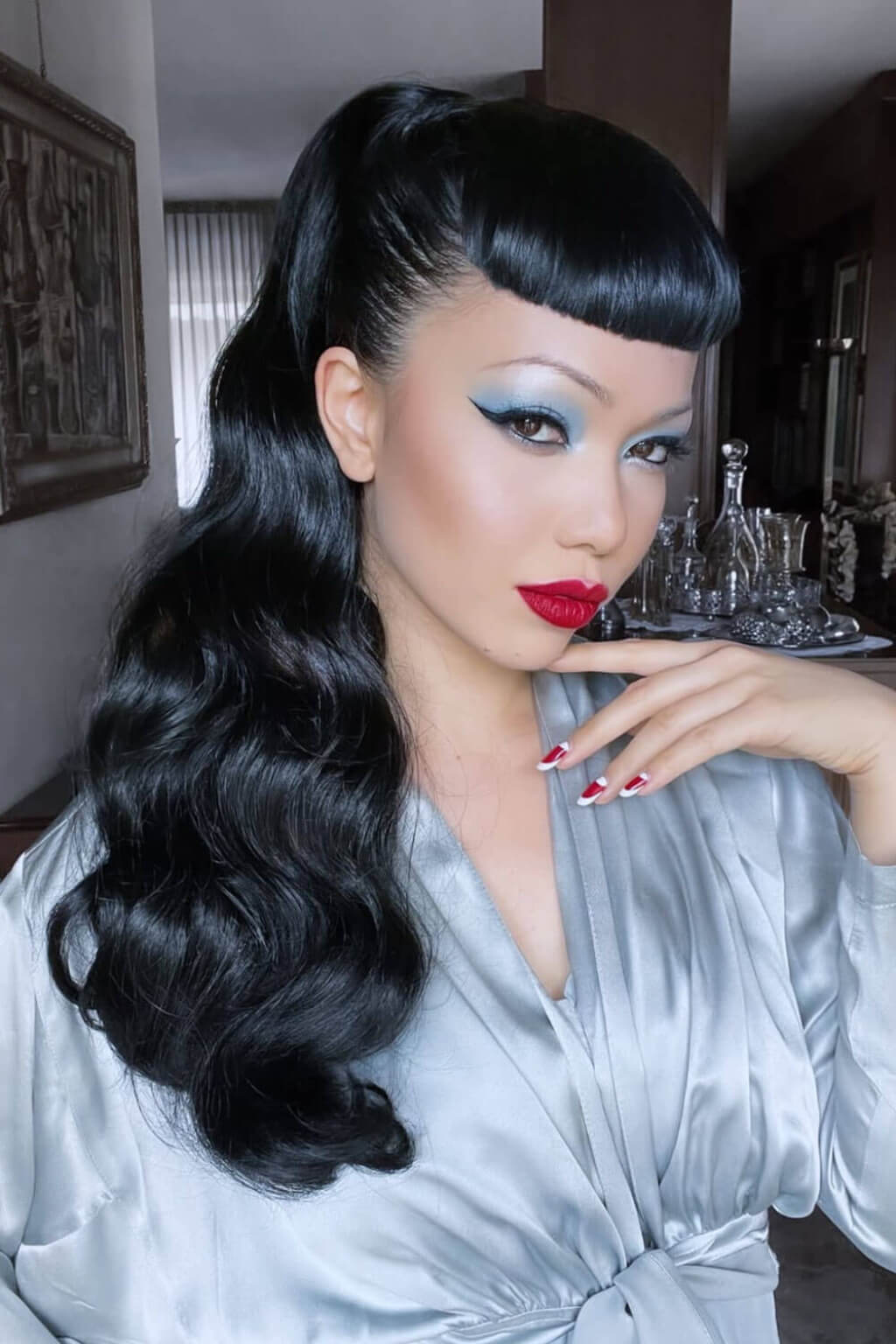 A woman with a retro 1950s style, wearing a black wavy wig with a high ponytail and thick front bangs, a light grey satin robe, and red lipstick. Long ponytail with vintage waves: Kitrysha jet black 1