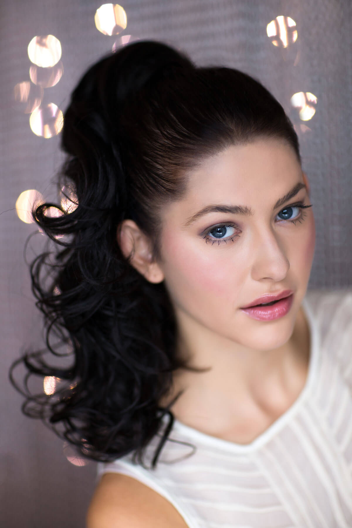 A woman wearing a curly ponytail extension hairpiece with loose, layered curls. She is seen from the shoulders up, with a backdrop featuring bokeh lights. Curly ponytail extension clip-on hairpiece: Tulisa