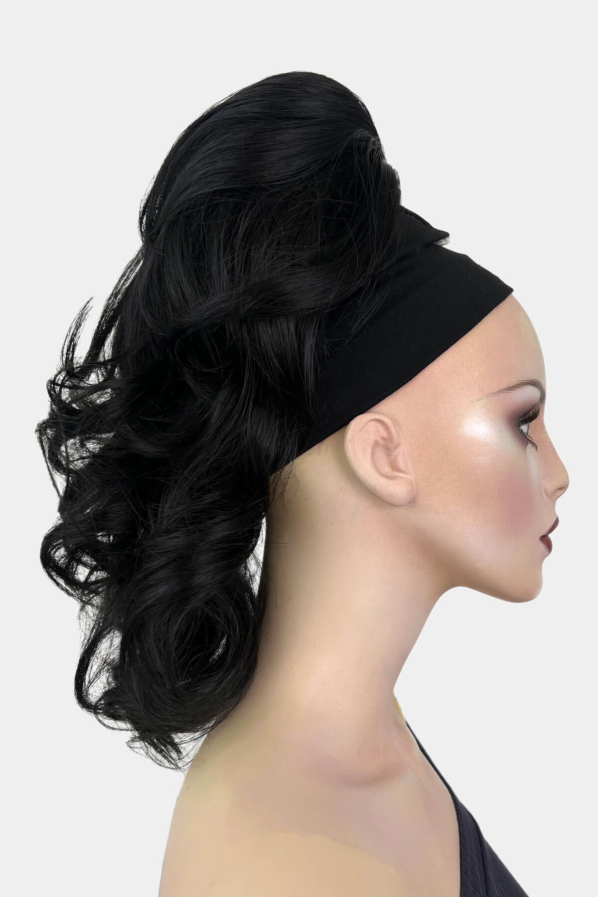 A mannequin wearing a curly ponytail extension hairpiece with loose, layered curls. It is seen from the shoulders up, with a backdrop featuring bokeh lights. Curly ponytail extension clip-on hairpiece: Tulisa black 1B