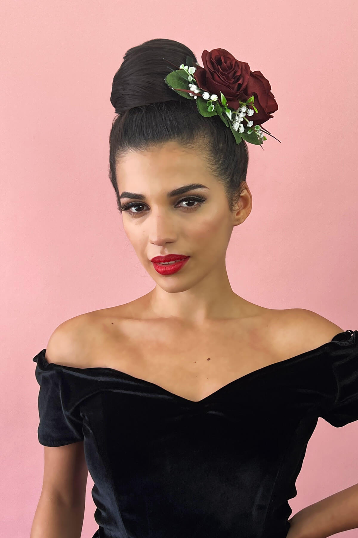Woman with flowers in her hair wearing a black off-shoulder top against a pink background.