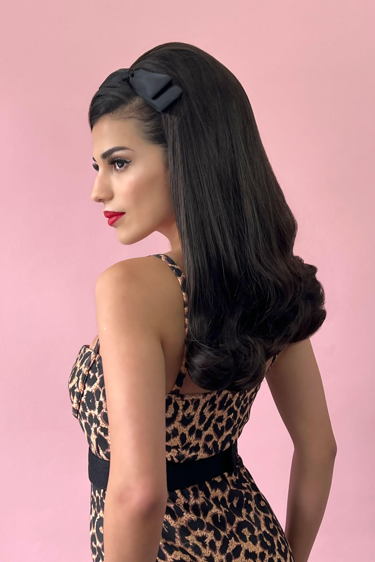 Woman with long, wavy hair wearing a black headband against a pink background. 1960s Retro high crown, half wig hairpiece, long with hair flip: Priscilla chestnut ginger