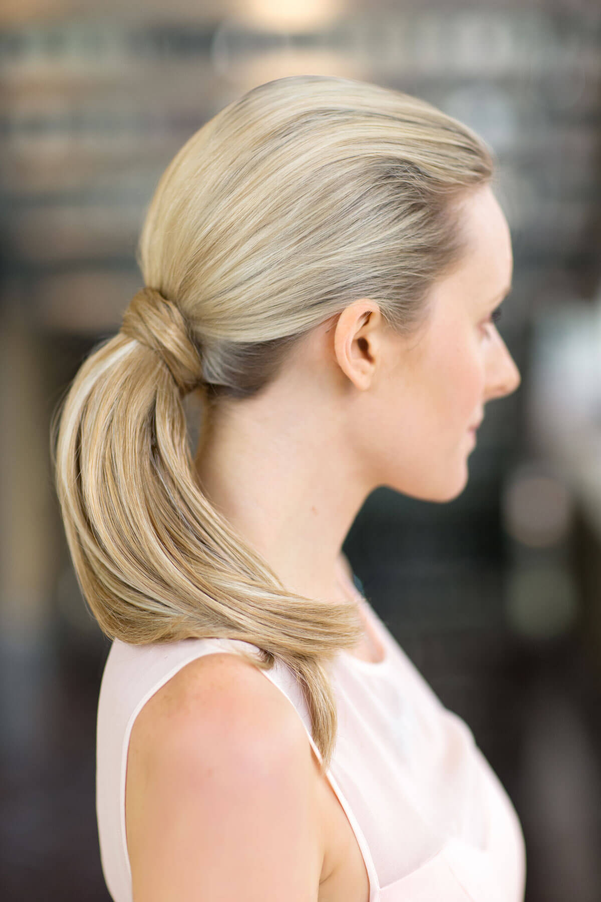 Woman with blonde hair styled in a ponytail against a blurred background. Sleek ponytail extension hairpiece, Flexihair: Jasmin