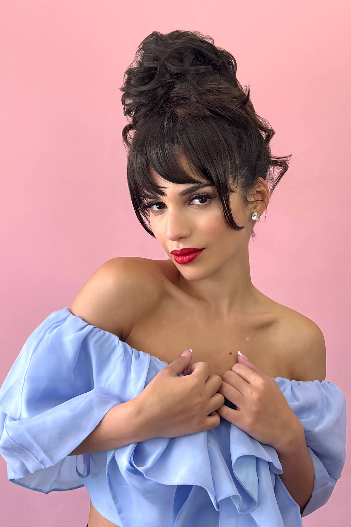 Woman with styled hair and red lipstick wearing a light blue off-shoulder top against a pink background. Scrunchie bun hairpiece, easy updo messy bun: Betsy ginger