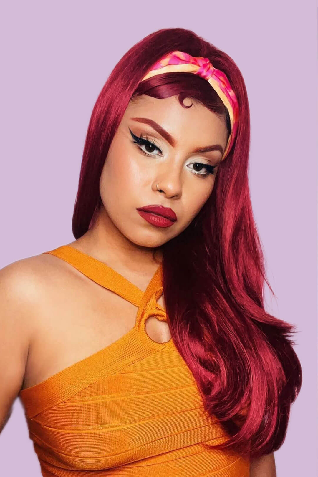 Woman with vibrant red hair and orange dress against a light purple background. 1960s style hair, high crown long straight and layered half wig: Juliette wine red 39