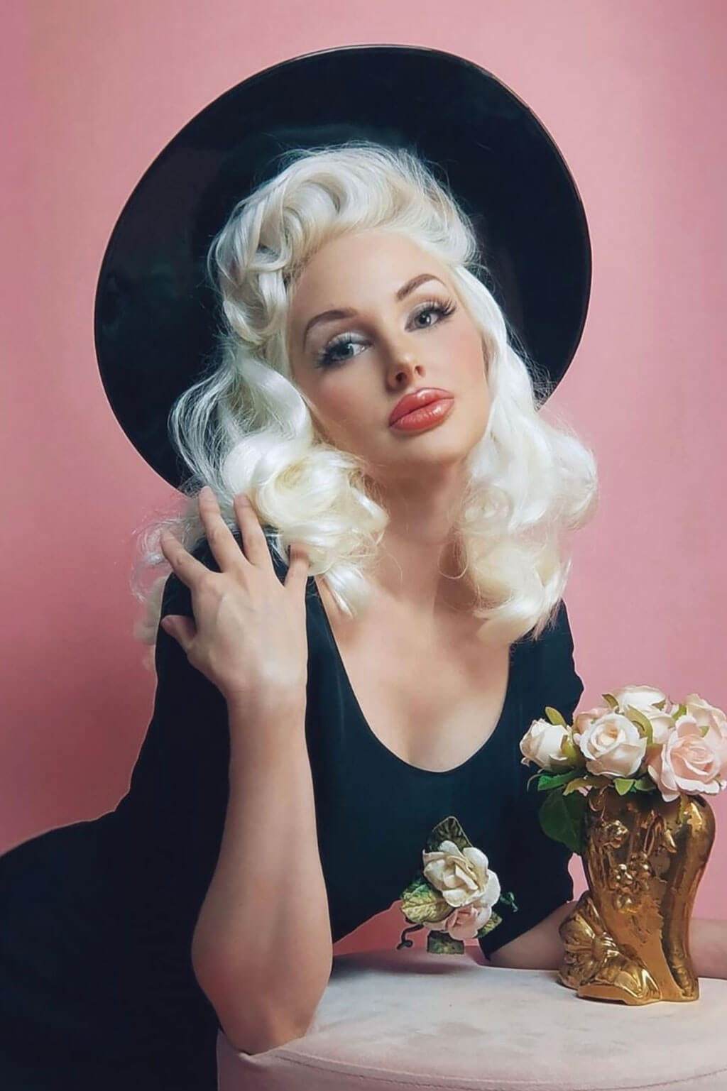 A person with shoulder-length black waves wearing a black sleeveless dress against a pink background.  1950s style shoulder length half wig hairpiece with beautiful vintage waves: Onyx platinum blonde 1001