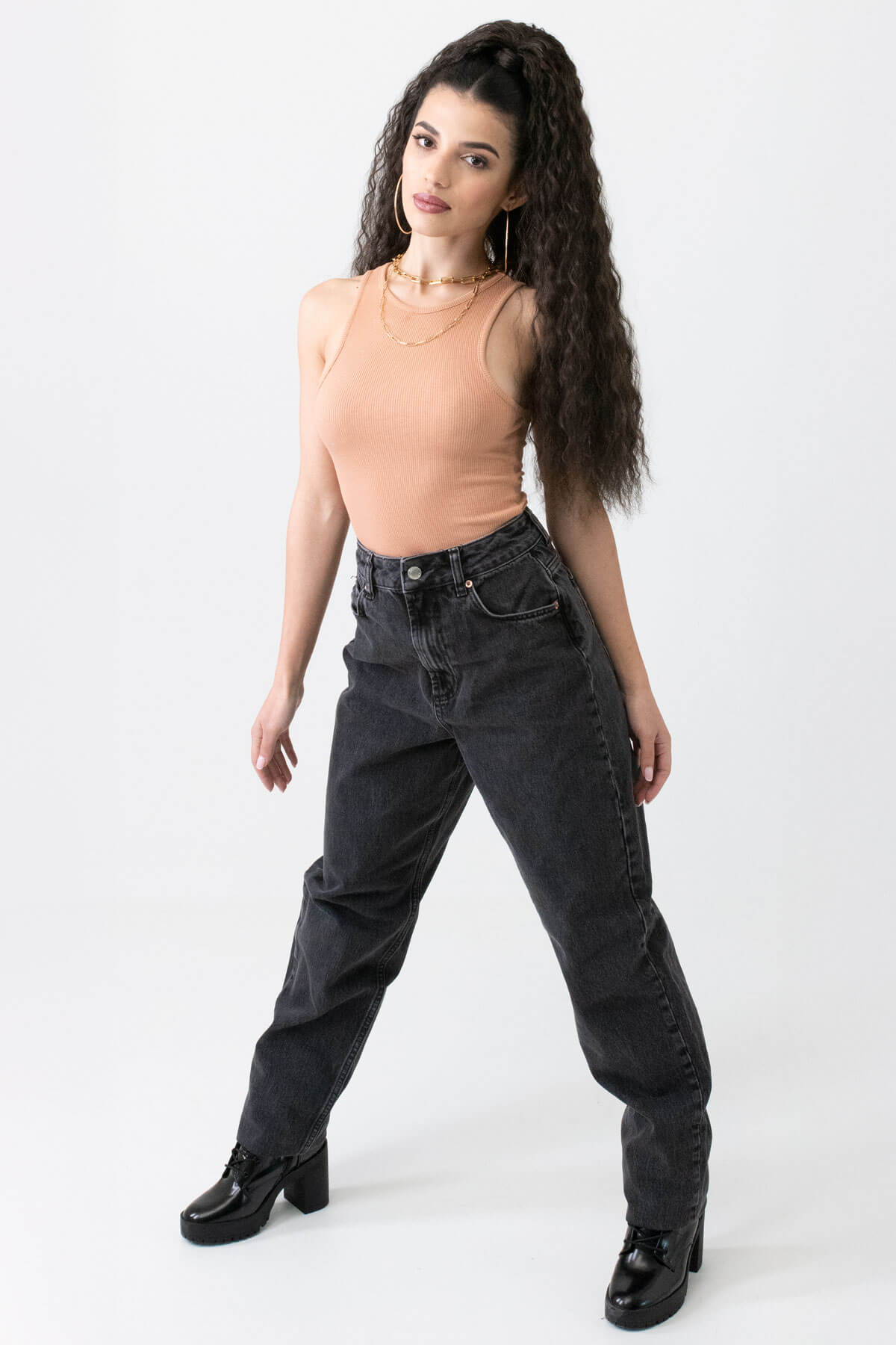 Woman wearing black jeans and a beige top on a white background.  Extra long ponytail extension with long loose spirals: Kaia dark brown 4