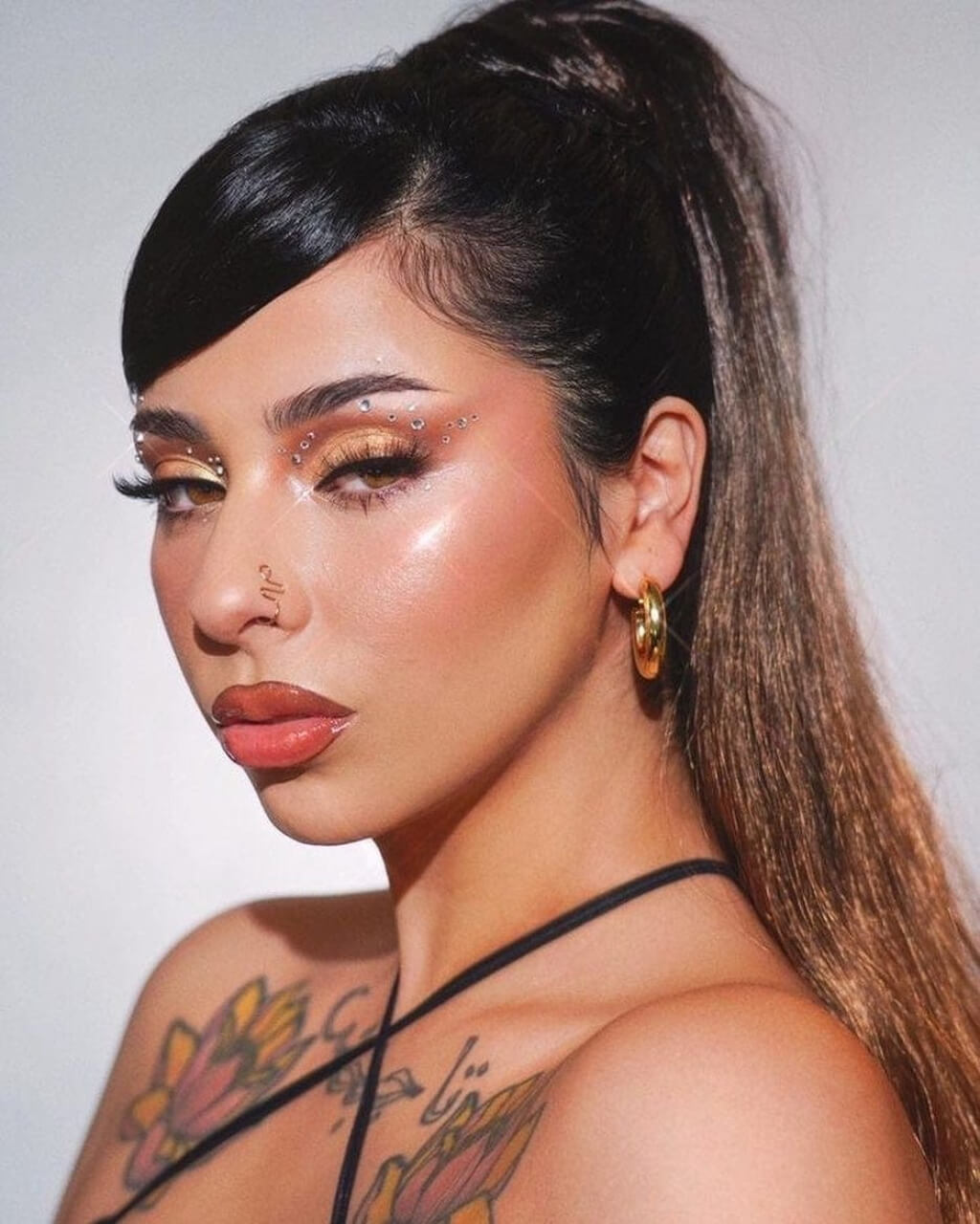 Woman with tattoos and gold makeup wearing a black halter top against a plain background. Long ponytail extension with soft waves: Sophie 4TN27
