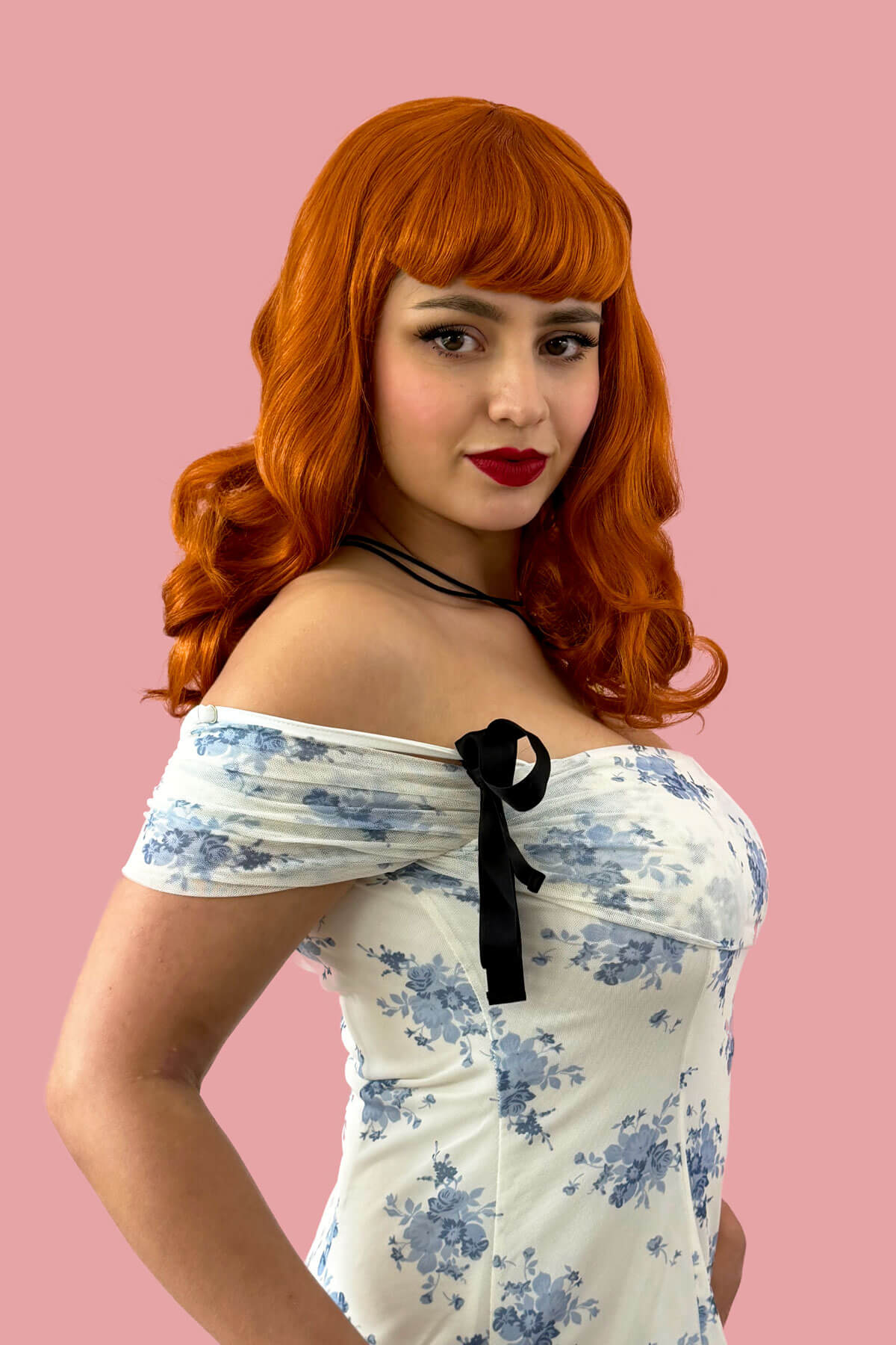 Woman with red hair and a floral off-shoulder dress against a pink background. Ginger pinup style wig, finger waved with short fringe, 1950s style: Sienna