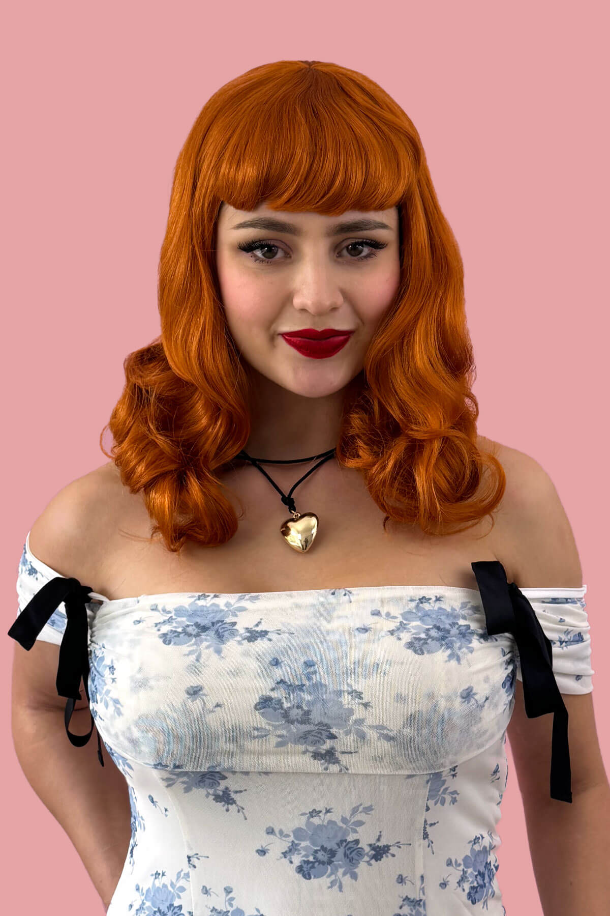Woman with red hair and a floral off-shoulder dress against a pink background. Ginger pinup style wig, finger waved with short fringe, 1950s style: Sienna