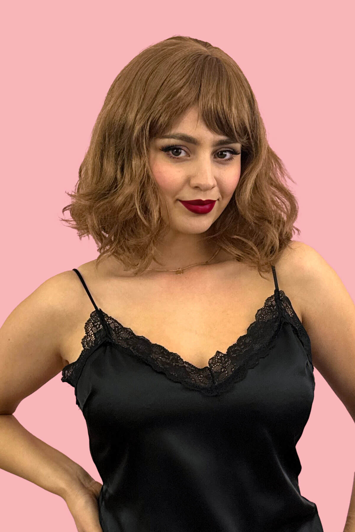 Woman wearing a black lace-trimmed top against a pink background. Brown long wavy bob (lob) wig with swept fringe: Clover