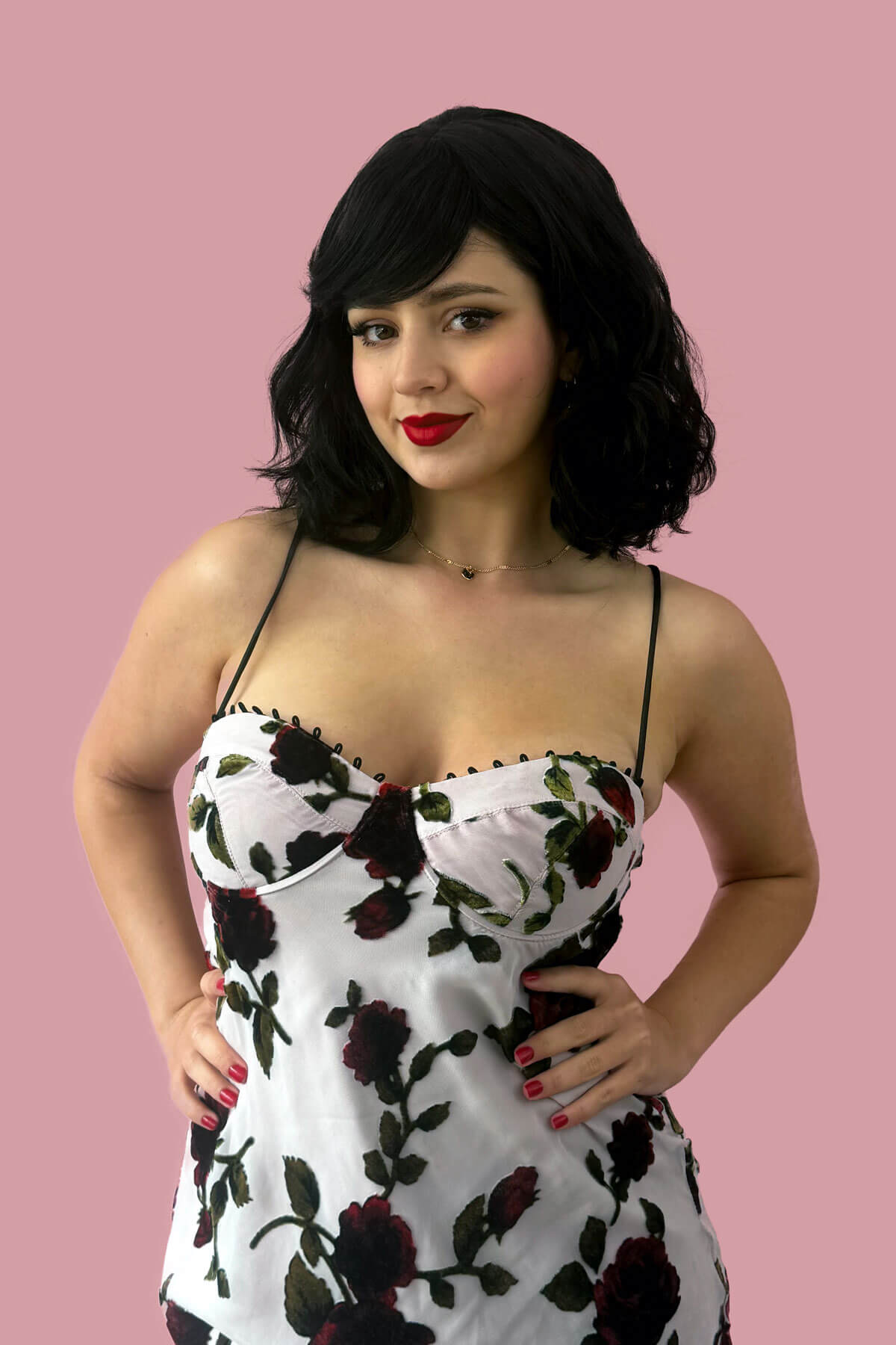 Woman wearing a floral dress against a pink background. Black long wavy bob (lob) wig with swept fringe: Eleena