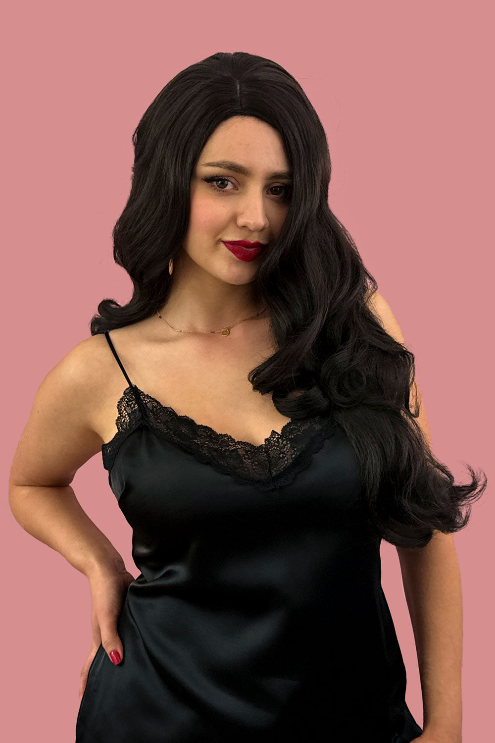 A person wearing a long brown wig with loose waves and curls, featuring a side-sweeping fringe. The individual is dressed in a black top and the background is pink.Long, brown wig, with wonderful loose waves and curls and a sweeping fringe: Leonie