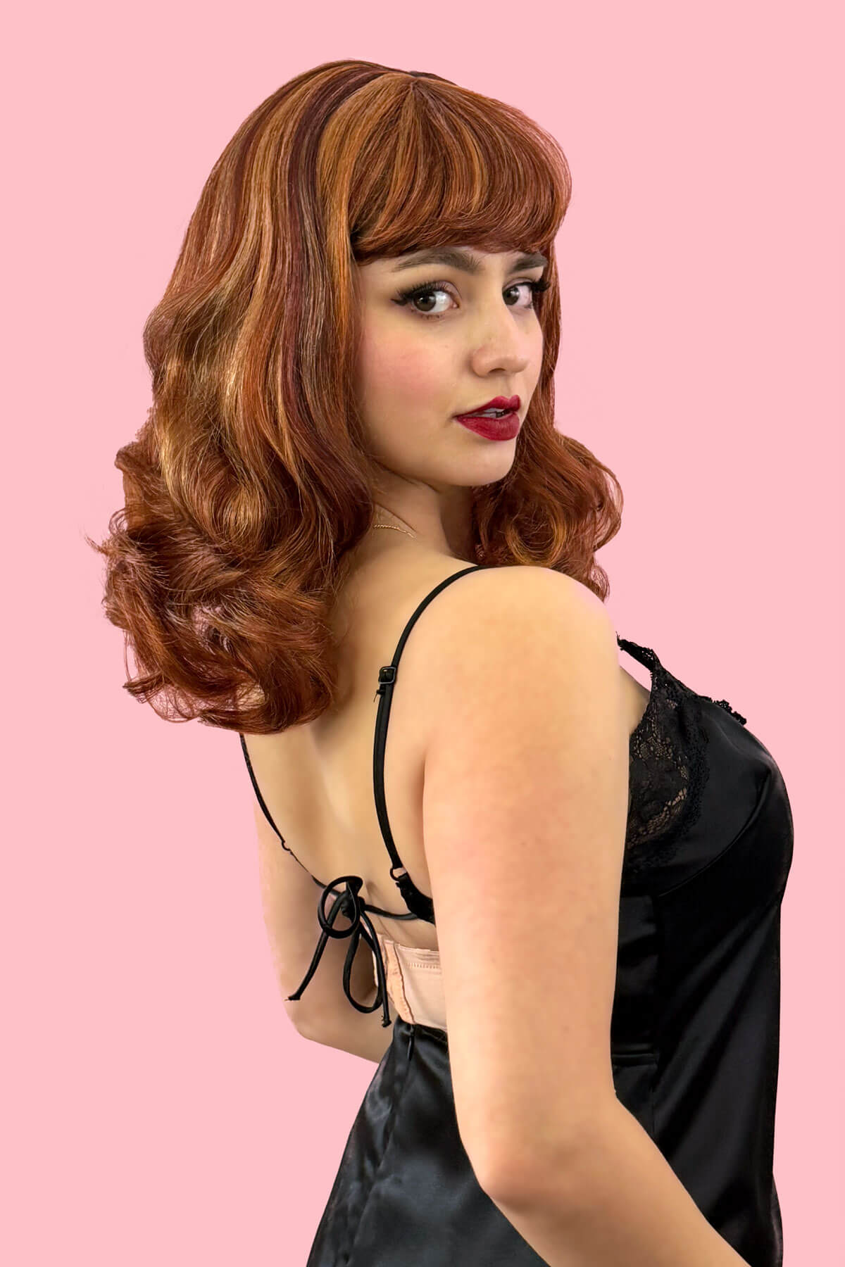Woman with a vintage-style wig and black lace top against a pink background. Auburn pinup style wig, finger waved with short fringe, 1950s style: Heidi