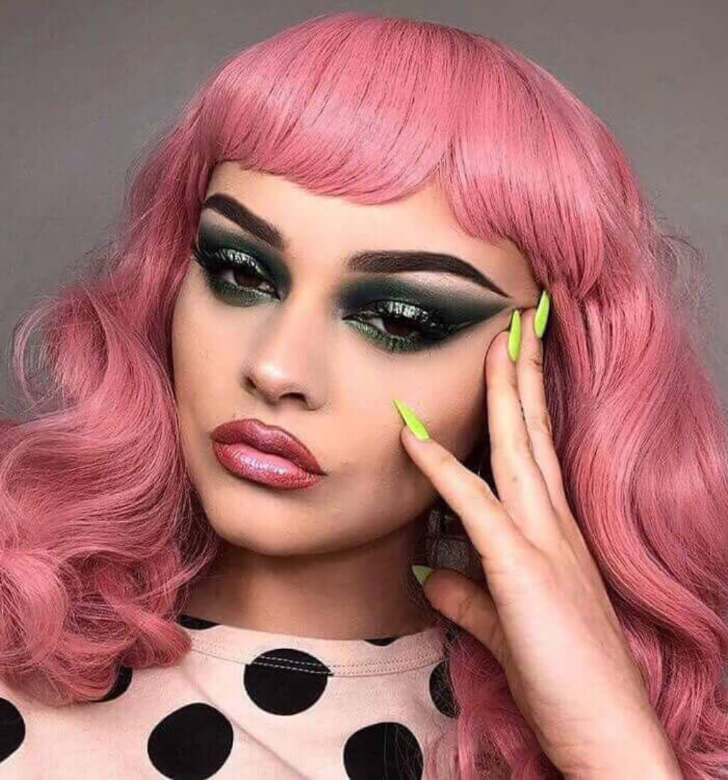 Person with pink hair and dramatic makeup touching their face, wearing a polka dot top. Pink pinup style wig, with finger waves and a short fringe, 1950s style: Stevie
