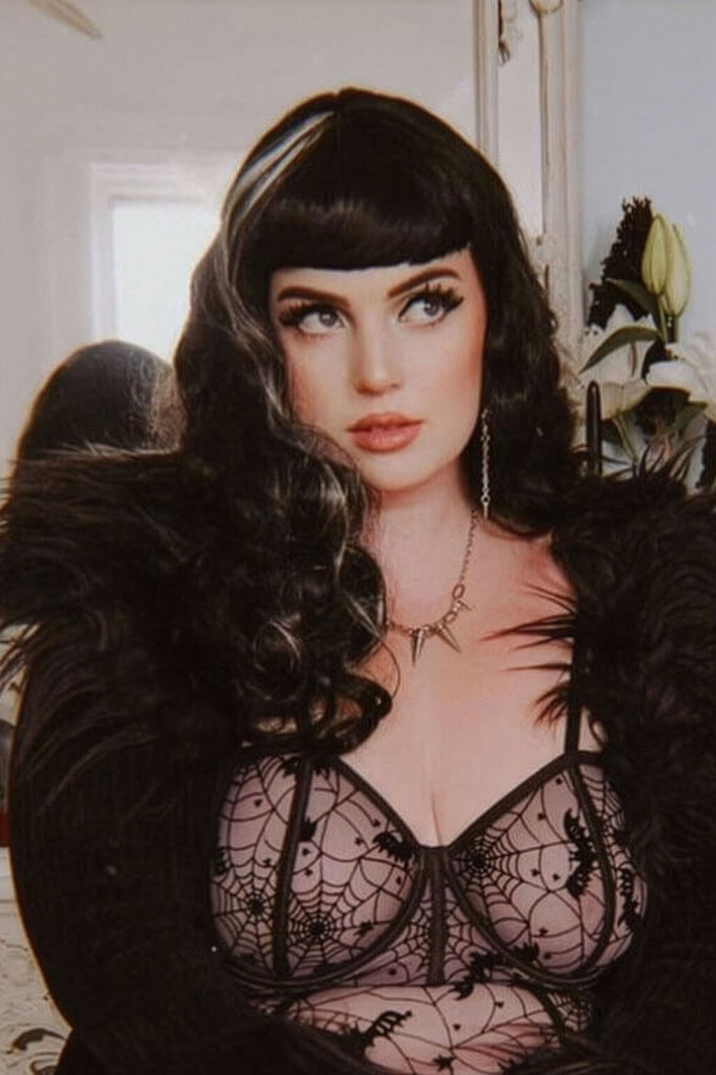 Woman wearing a black lace lingerie set with a feathered black jacket indoors. Long black 1950s pin up style wig with platinum streak and retro fringe: Michelle