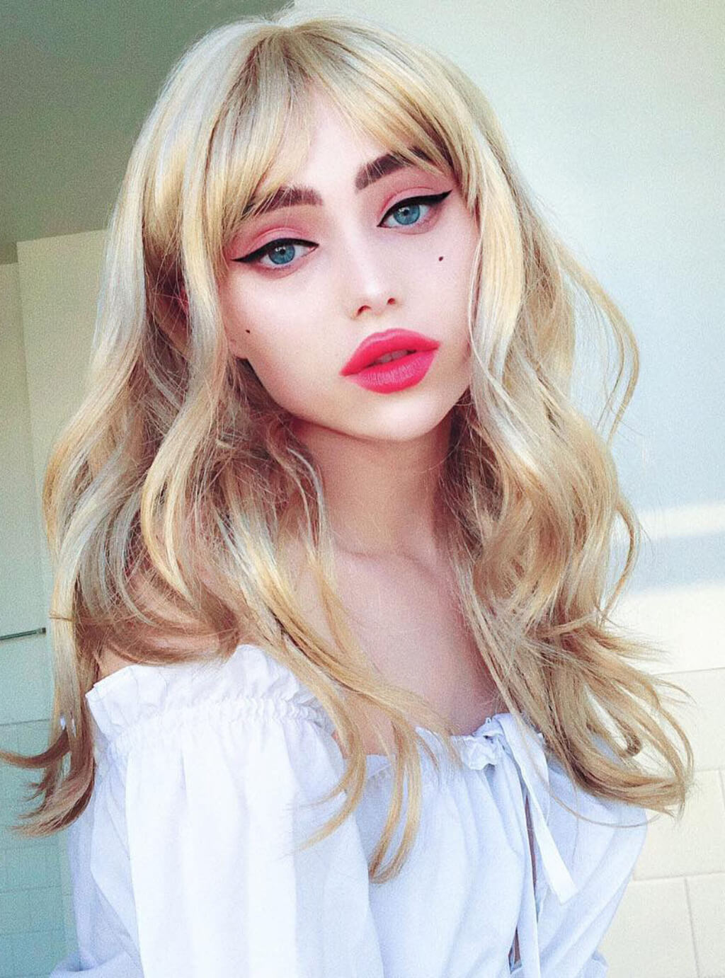 Person with blonde wig and white blouse against a light background. Long ash blonde wig with gentle waves: Harriet
