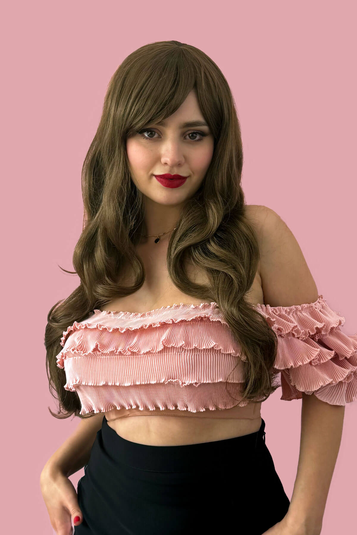 Woman wearing a pink ruffled top against a pink background. Long wavy light brown wig with vintage-style straight fringe: Maya
