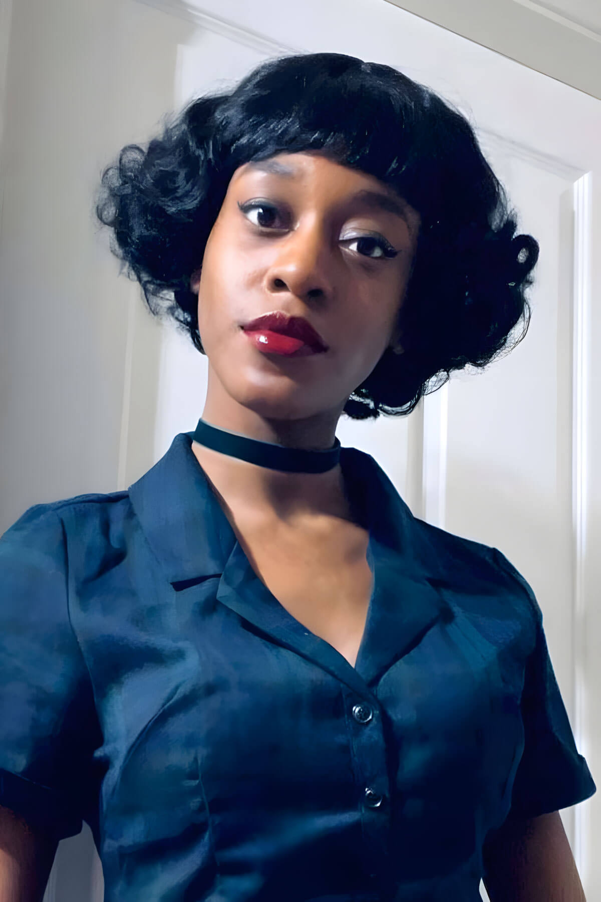 Woman wearing a teal dress with a black choker in front of a white door. A short black vintage style wig with finger waves: Marlene