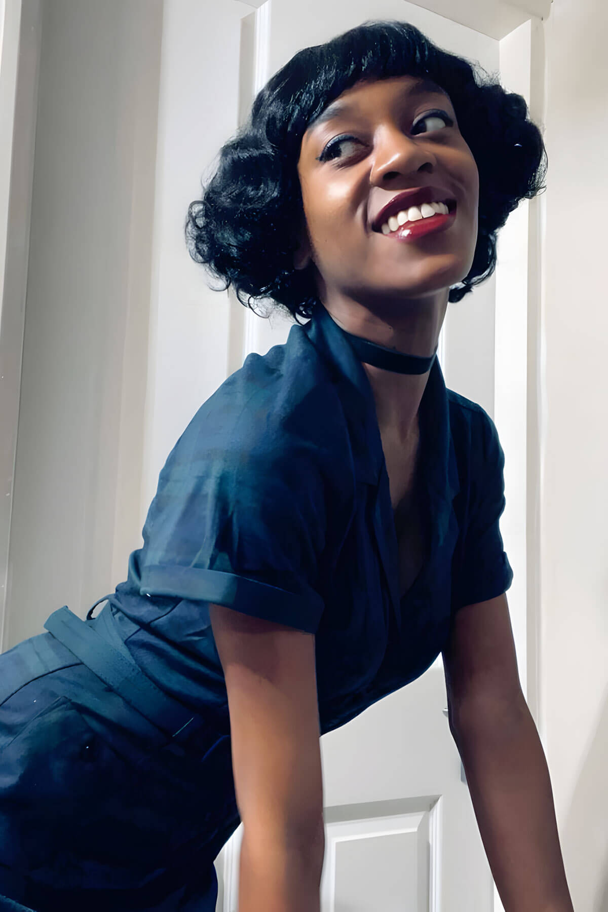 Woman in a blue dress smiling in front of a white door. A short black vintage style wig with finger waves: Marlene