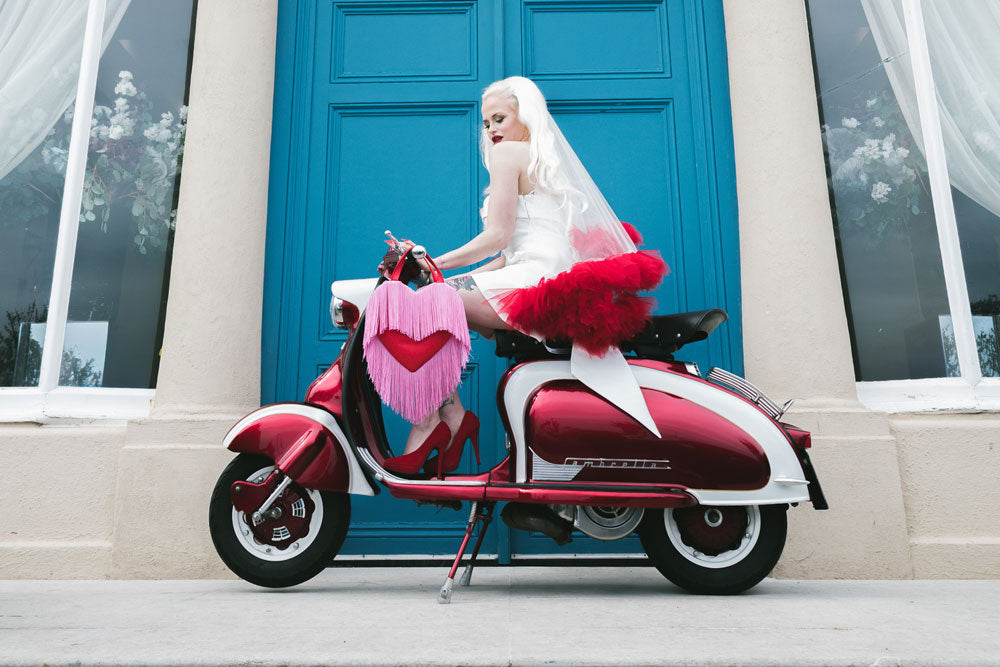 Person in a white dress with red accents sitting on a red scooter in front of a blue door with long blonde hair.