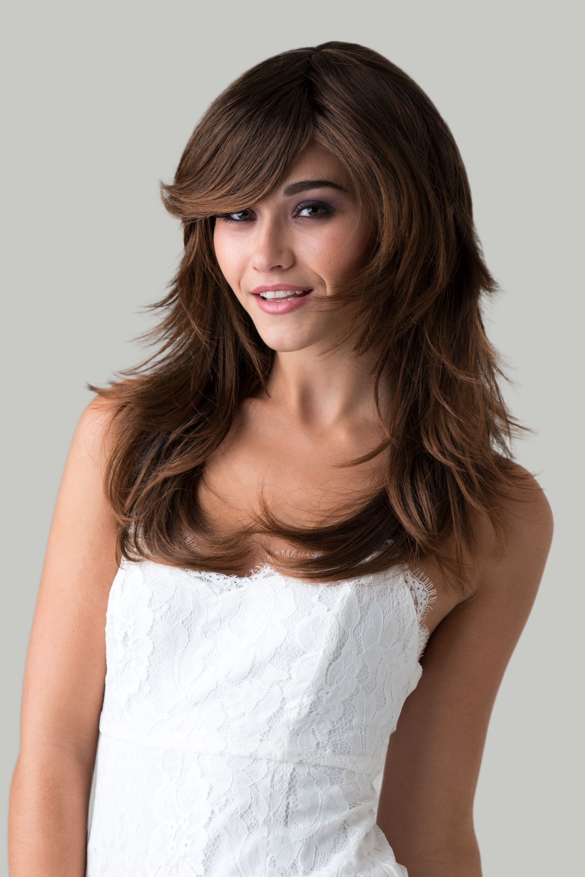Woman with long brown hair wearing a white dress against a gray background. Long warm brown wig, layered, flicked tips: Selina