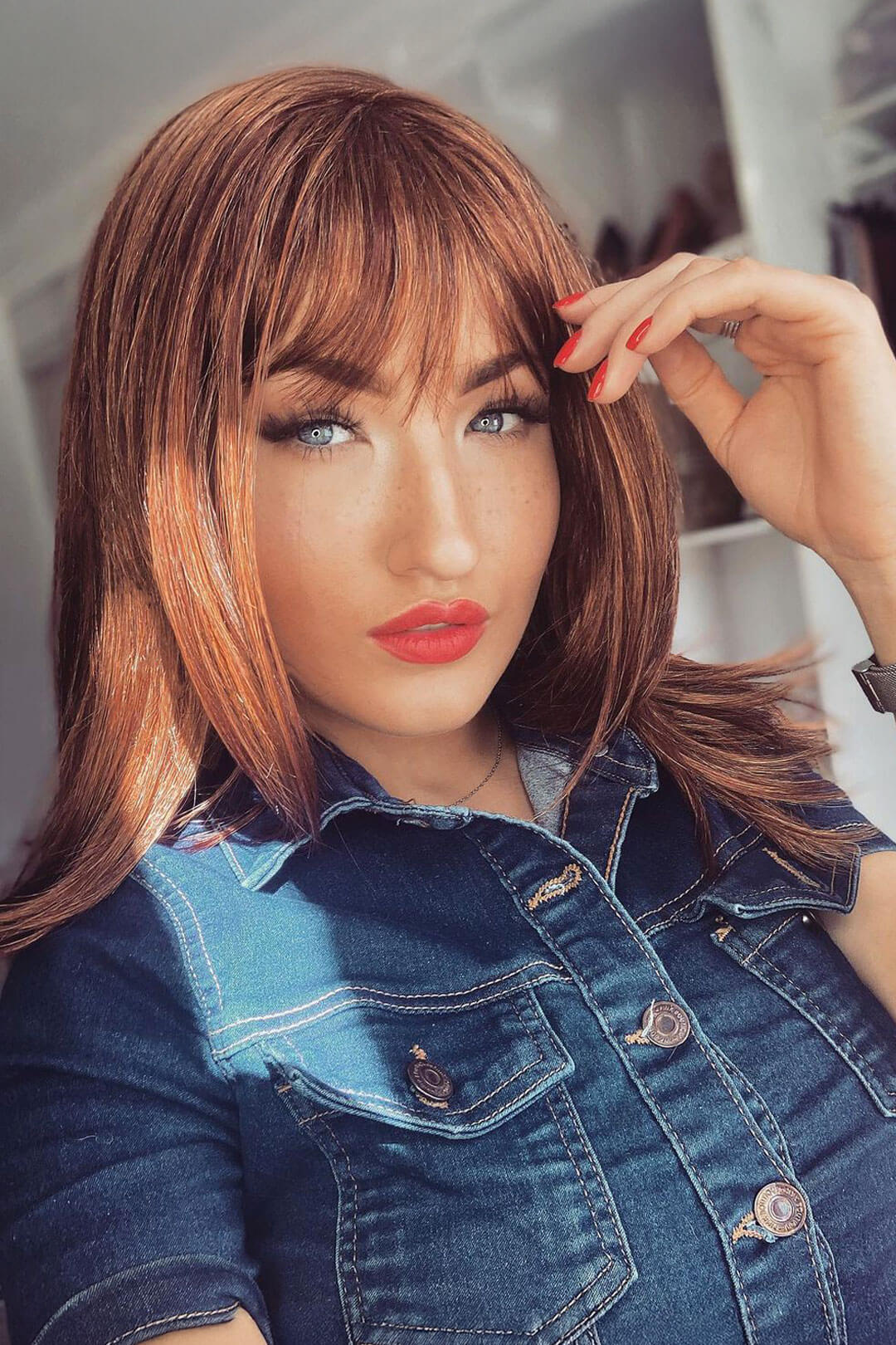 Woman with styled auburn hair and denim jacket posing indoors. Gingery auburn wig with blonde highlights face frame style: Geri