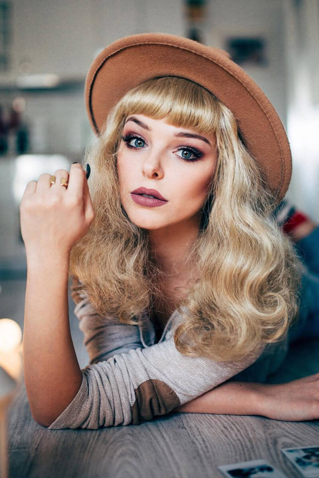 A woman with long wavy blonde hair and bangs wears a brown wide-brim hat and a gray top, leaning on a table. Blonde pinup wig, curled with short fringe, 1950s style: Cora blonde 24H613
