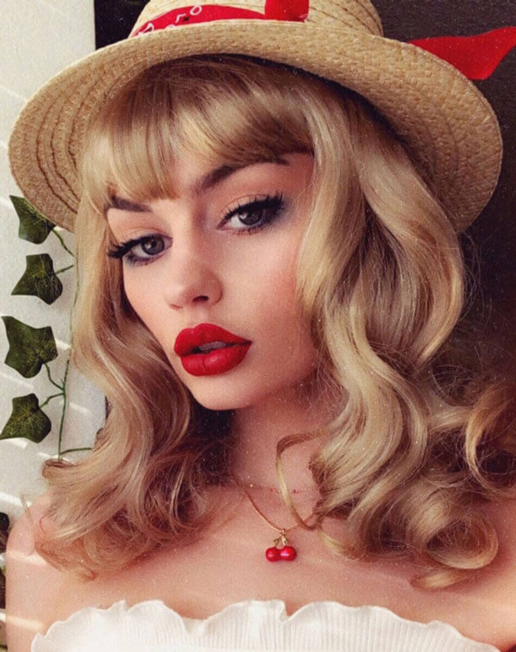Woman with blonde hair, red lipstick, and a straw hat with a red bandana. Blonde pinup wig, curled with short fringe, 1950s style: Cora blonde 24H613