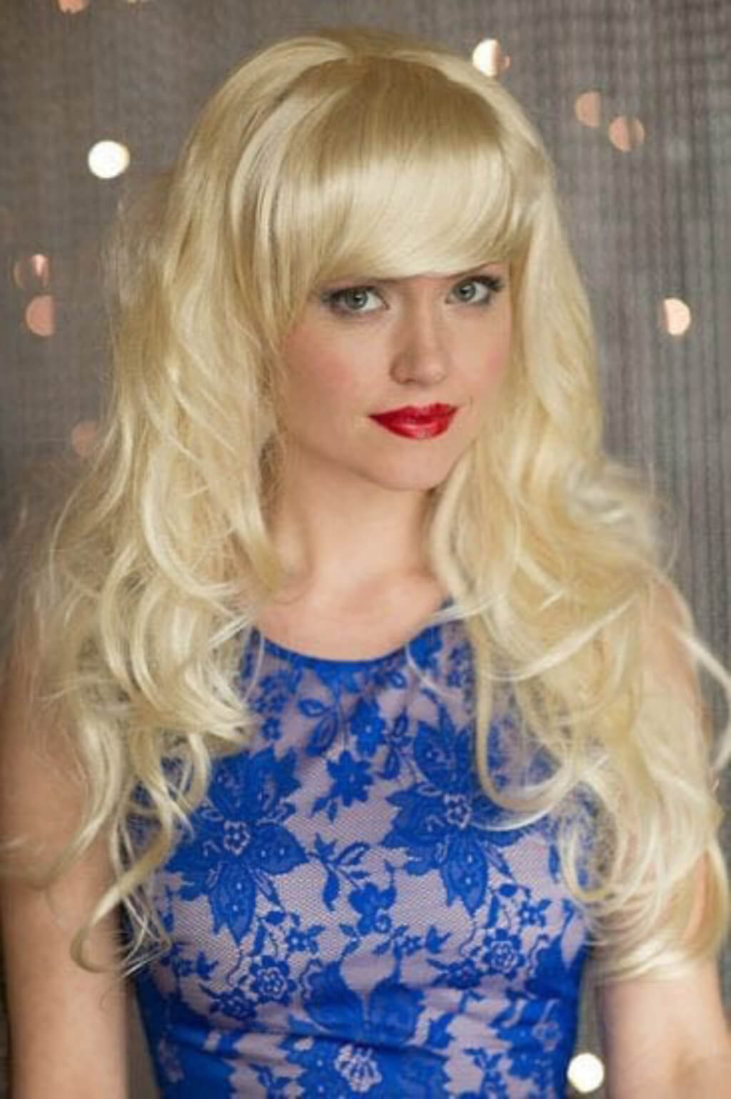 Woman with blonde wig and red lipstick wearing a blue lace dress against a neutral background. Clip-in fringe hairpiece, curved, side-sweeping/clip-in bangs: Eliana blonde 613 
