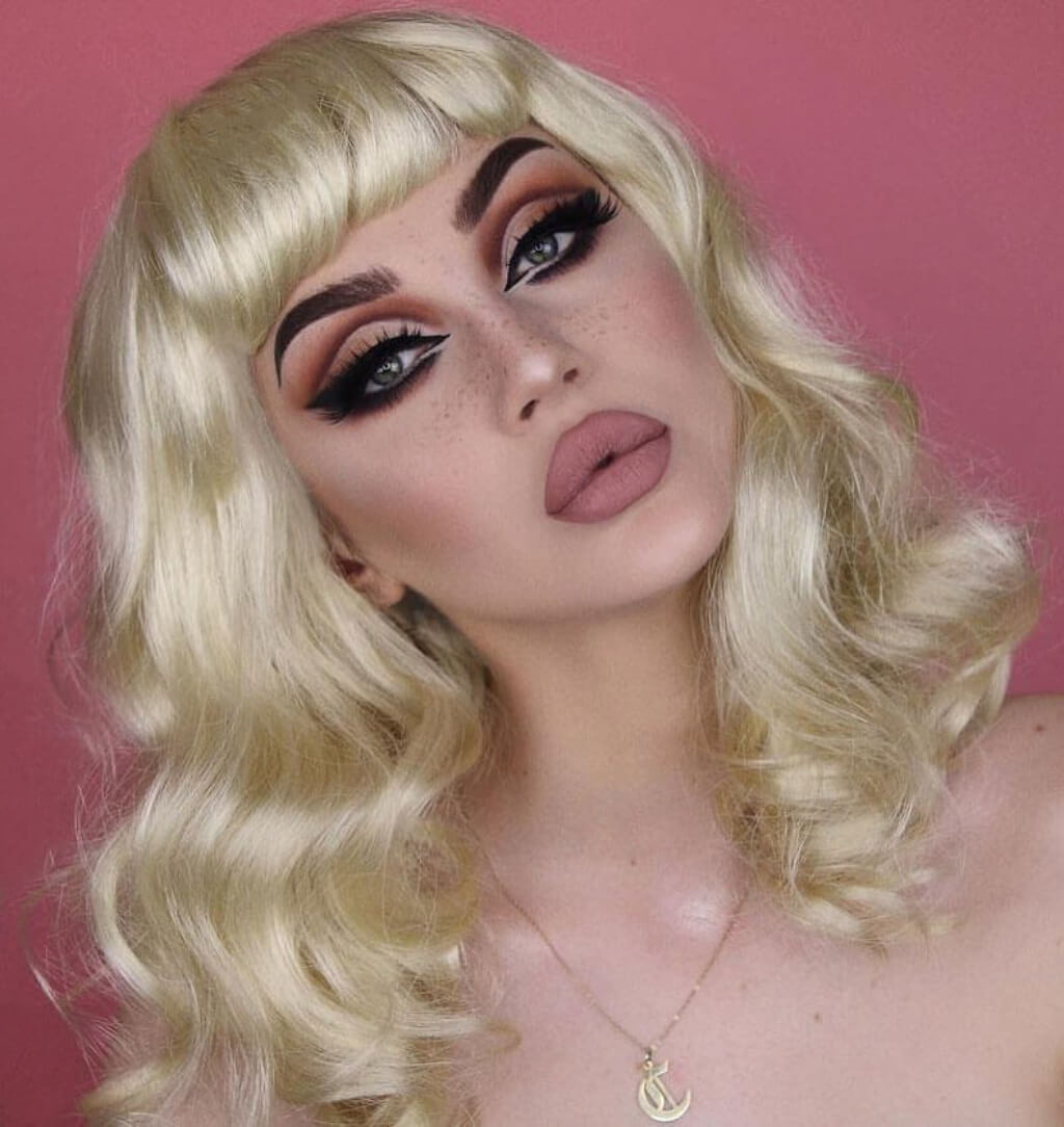 Person with blonde wig and dramatic makeup against a pink background. Blonde pinup wig, curled with short fringe, 1950s style: Cora creamy blonde 613