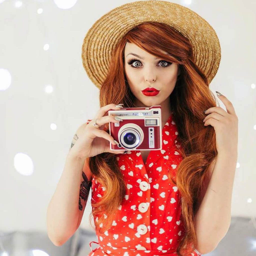 A woman with long red hair wearing a straw hat and a red dress with white hearts, holding a retro red and silver camera in front of her face against a bright, softly blurred background. Red & auburn wig with blonde highlights, long: Freya