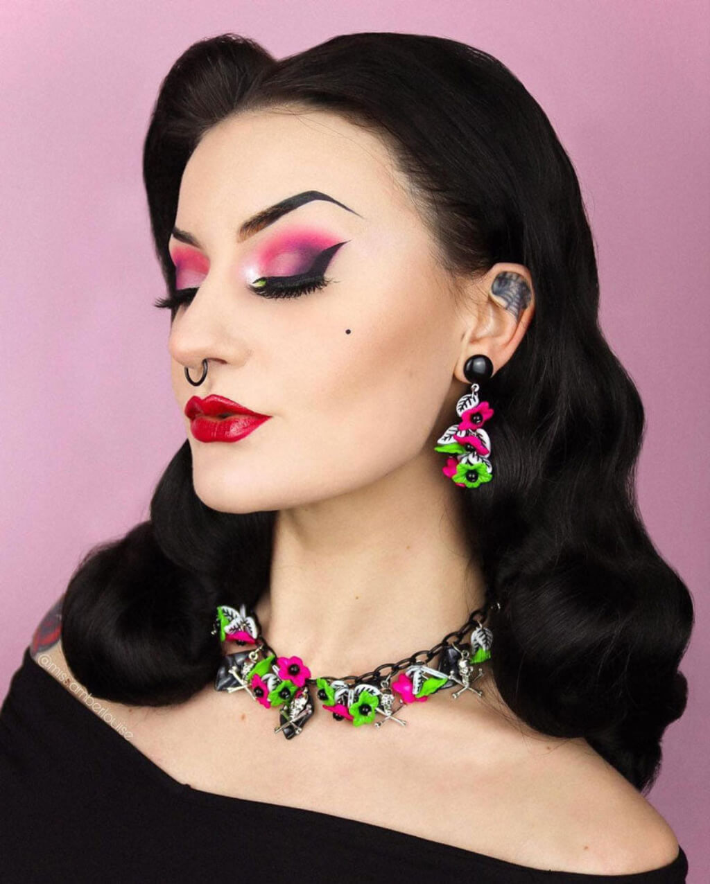 Woman with vintage-style makeup and hair, wearing colourful jewellery against a pink background. Black soft glamour wig with long marcel waves: Vivienne