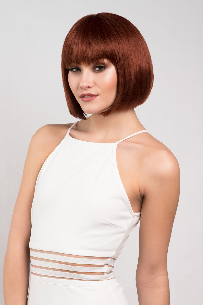 A woman wearing a copper-red bob wig with a heavy fringe, styled to fall just to the chin, modeled in a white tank top. Short copper-red/ginger bob wig: Minnie